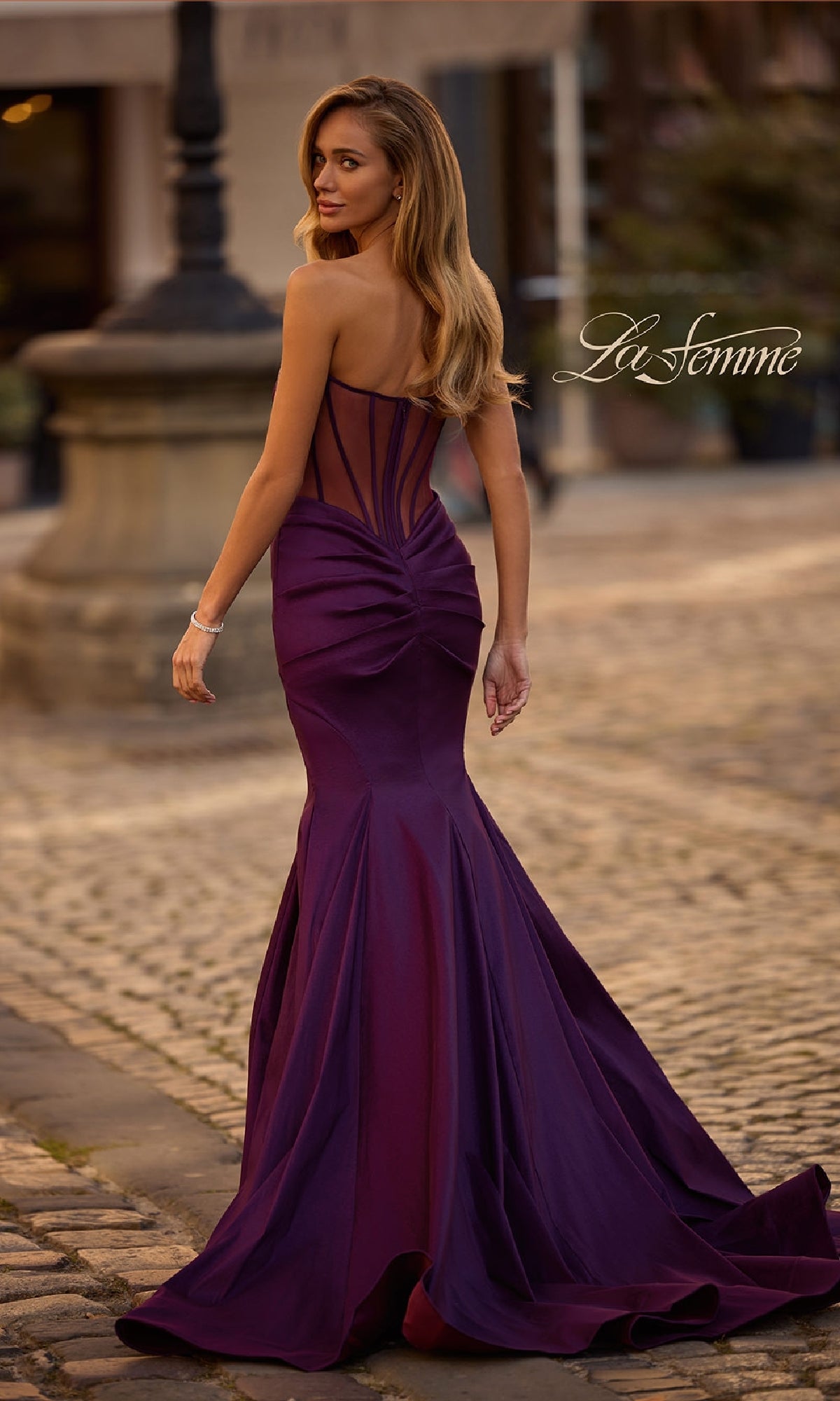 A woman in the Strapless Long Mermaid Prom Dress: La Femme 32791 stands on a cobblestone street, facing away, her fitted purple gown with a sheer back and elegant mermaid silhouette exuding sophistication.
